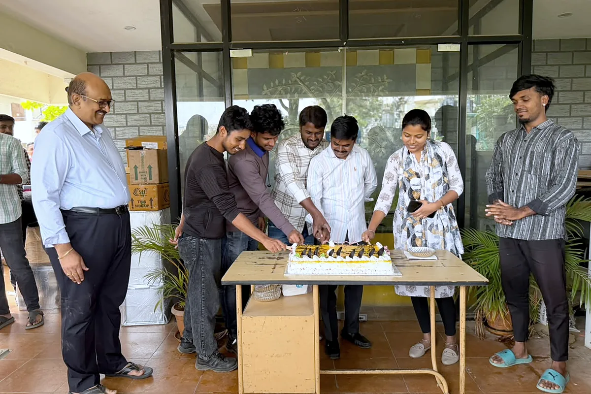 The team crowded round the porch table cutting a birthday cake, colleagues watching from either side.