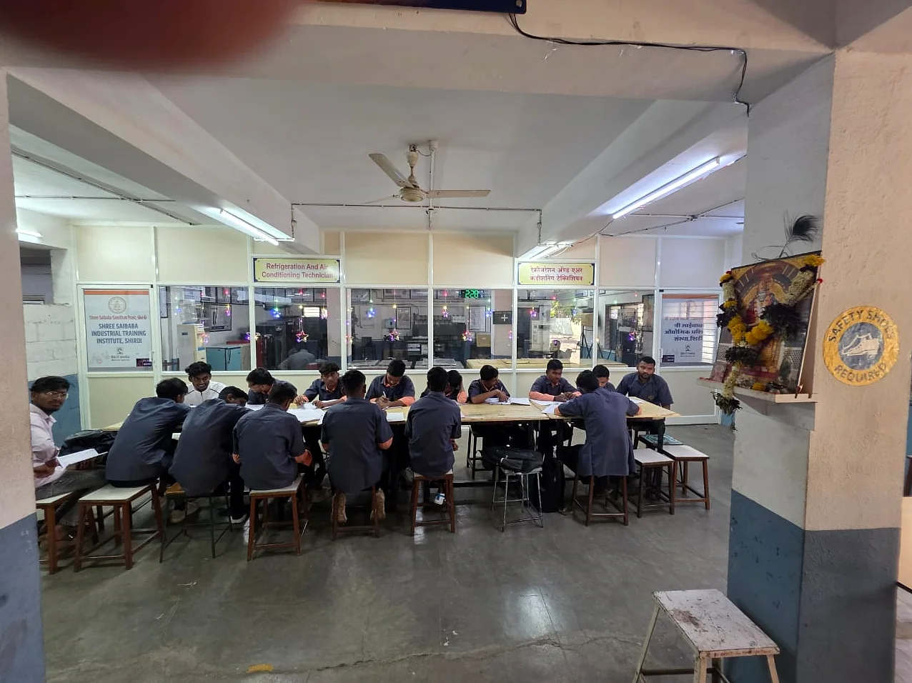 A classroom of students at shared benches, working under ceiling fans.