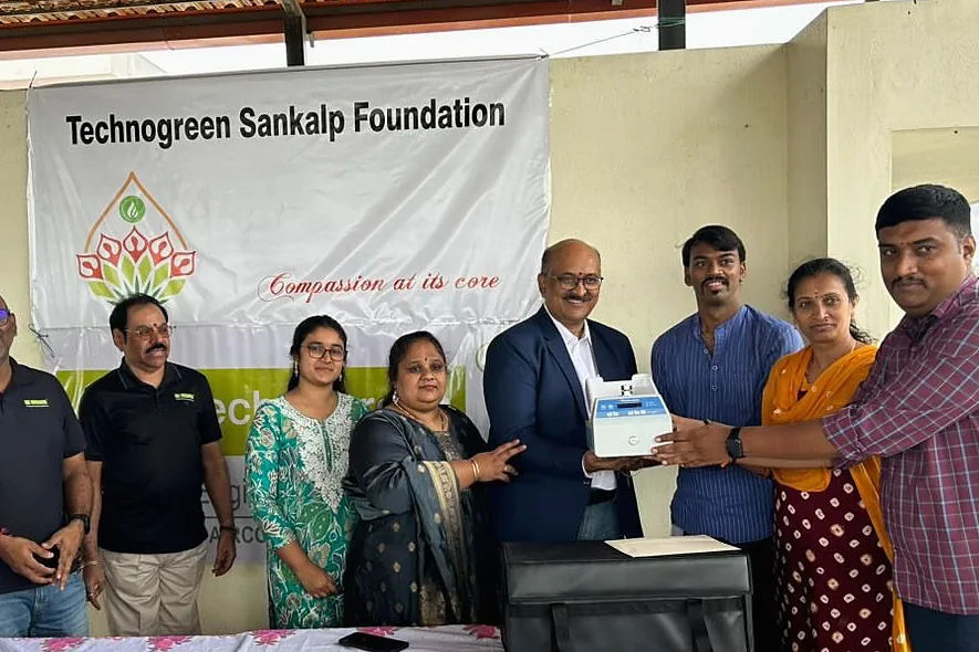 A boxed medical monitor handed across a table under a Technogreen Sankalp Foundation banner.