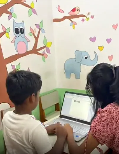 A child working at a laptop with a therapist, in a room painted with a tree and animals.