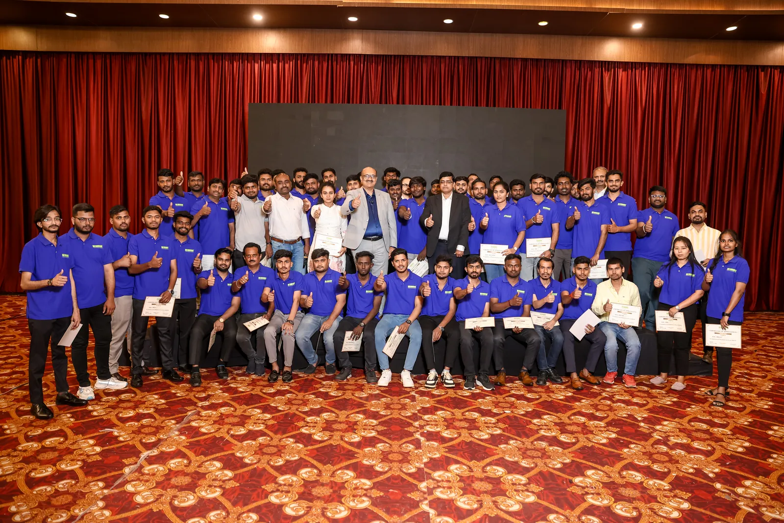 The team in matching blue T-shirts lined up across a banquet hall stage, holding certificates.