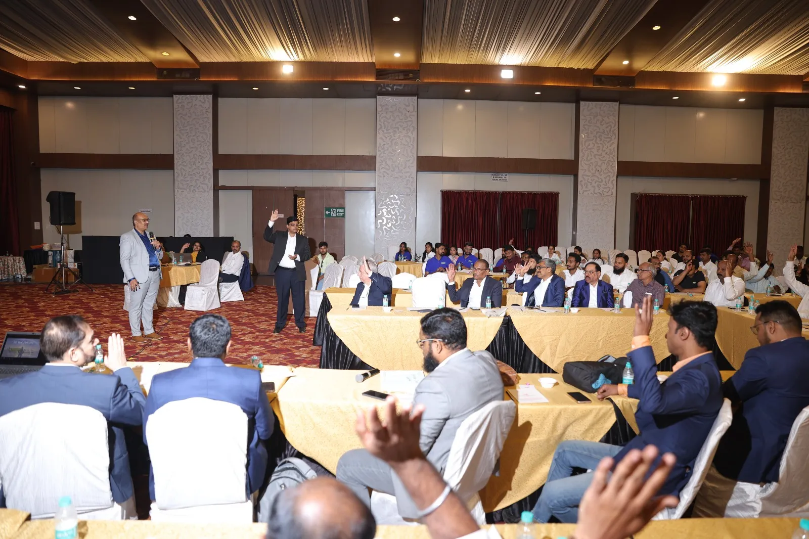 A packed session at long tables, hands raised in the front rows.