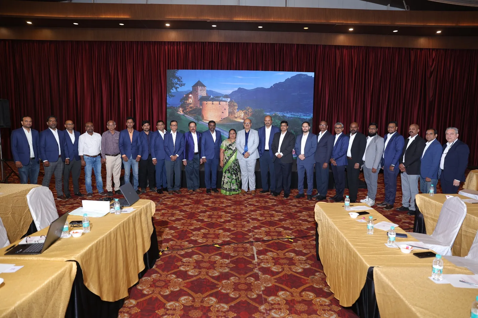 A line of managers in blazers standing in front of a projected slide between the meeting tables.