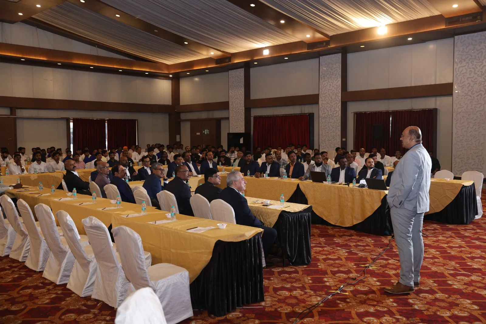A speaker addressing the room from the floor, rows of tables and chairs behind him.